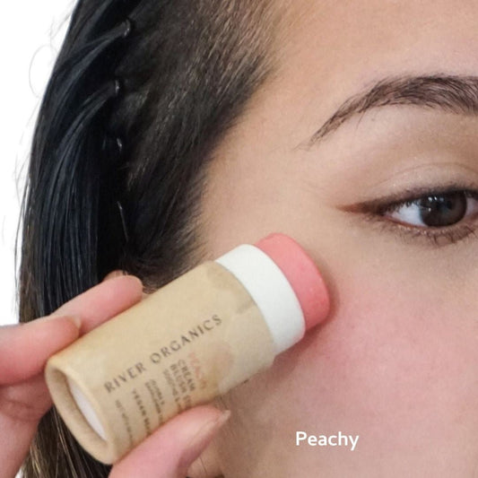 Someone applies a River Organics Set of 2 | Natural Cream Blush Sticks in peach to their cheek. The focus is on the stick and their skin, with "Peachy" displayed in the image.