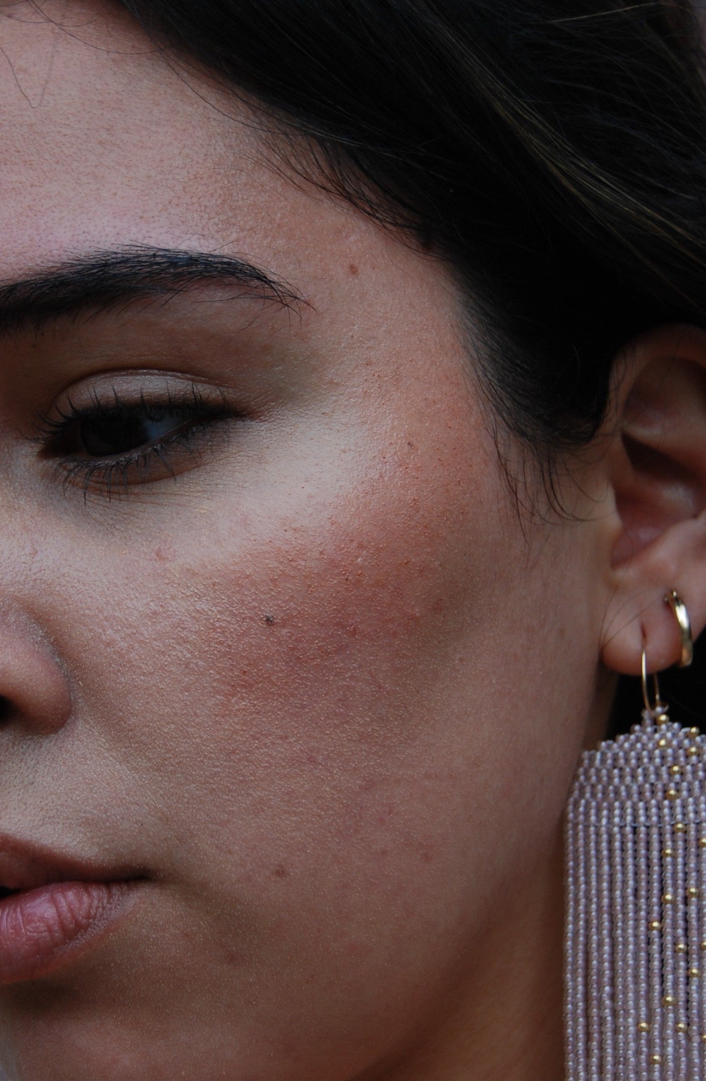 Close-up of a woman's face showcasing smooth skin with natural makeup, featuring River Organics Creamy Vegan Blush Stick in Pinch on her cheek and a beaded tassel earring. Her eyebrow, eye, lips, and ear are also visible.