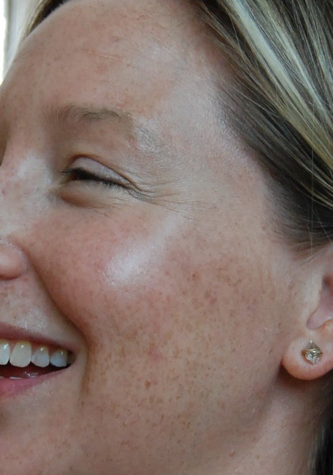 Close-up of a smiling woman with freckles and smooth skin, her right cheek glowing with River Organics’ Rose Quartz Cream Vegan Highlighter Stick. She has light brown hair with blonde highlights and wears a small earring.