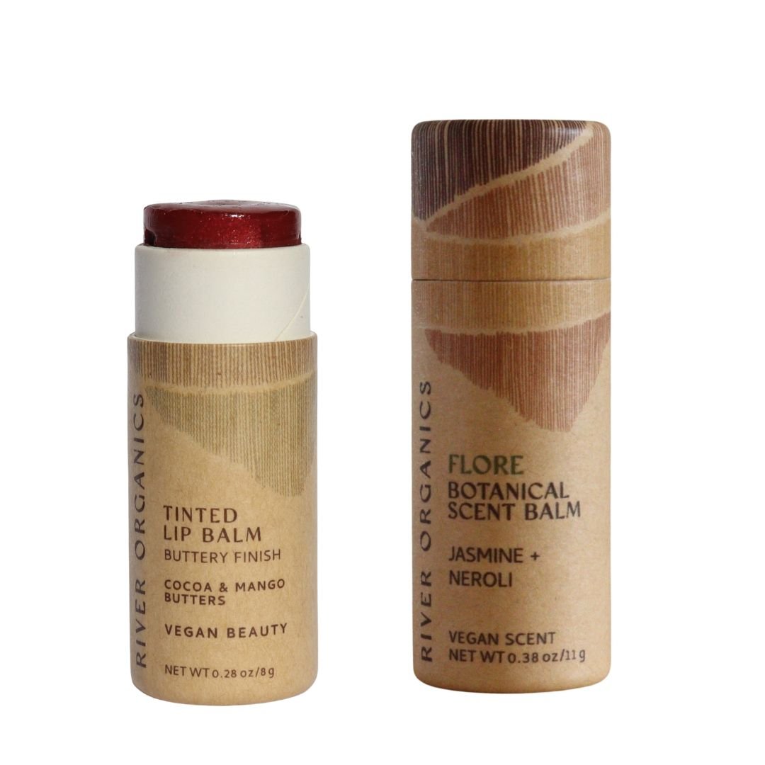 The River Organics Lip + Scent Balm Bundle features two cylindrical tubes: one vegan lip balm with the cap off to reveal a dark red shade, and one scent balm capped. Both have minimalist brown labels for your botanical ritual.