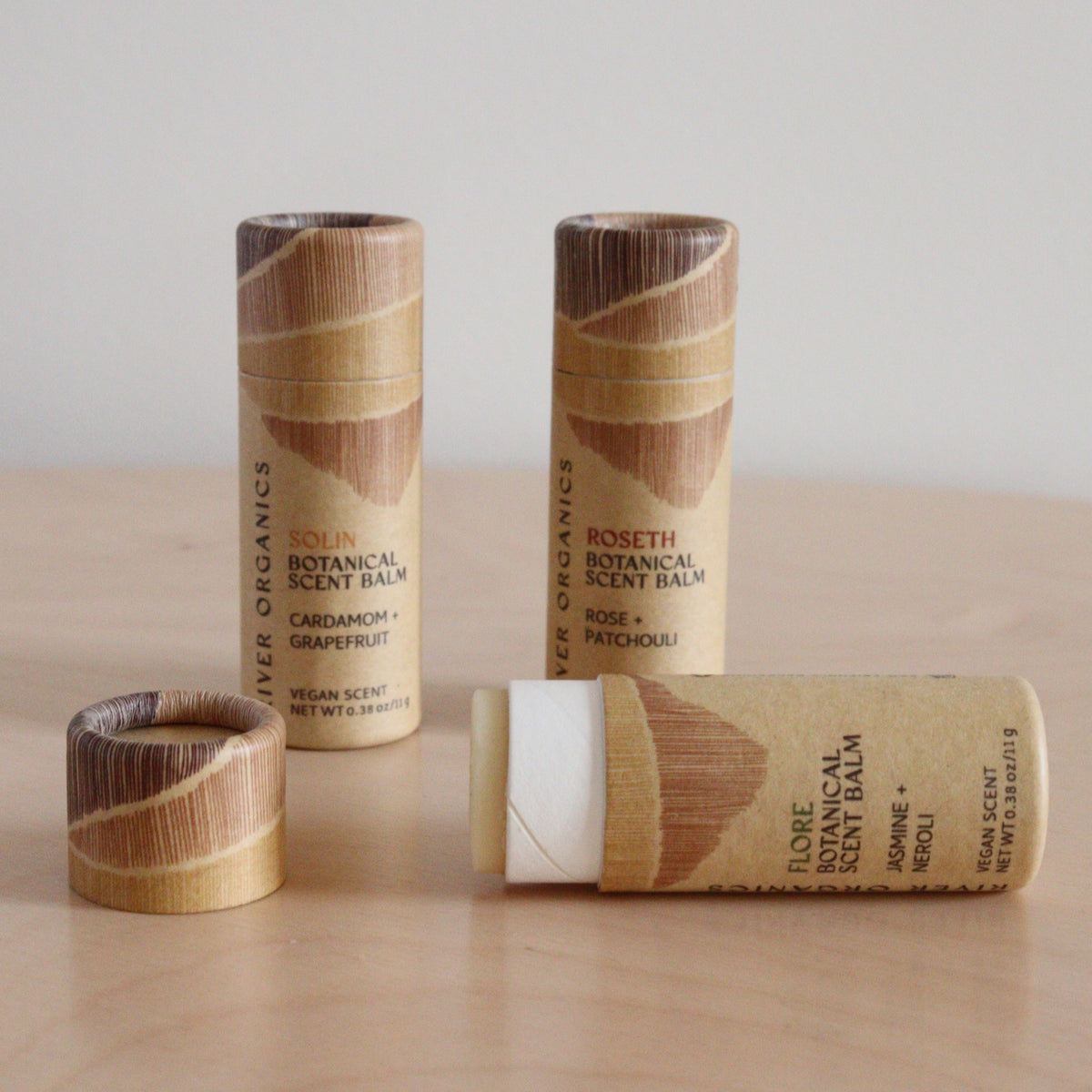 Four River Organics botanical scent balm tubes made with essential oils are displayed on a light wooden surface—three upright, one uncapped to show the solid perfume. Labels show different scents. Available as a set of 2, 3, or 4.