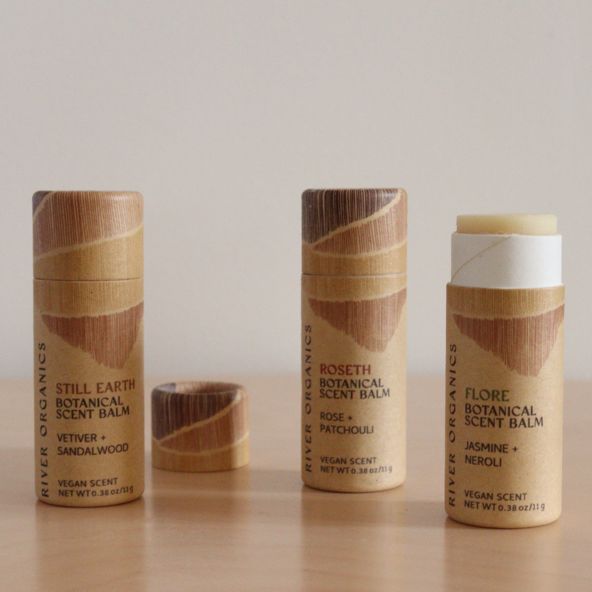 Three upright River Organics Botanical Scent Balms, made with essential oils and plant-based ingredients, are shown on a light surface. One balm’s cap is off to reveal the product inside. Labels display different scents. Available in sets of 2, 3, or 4.