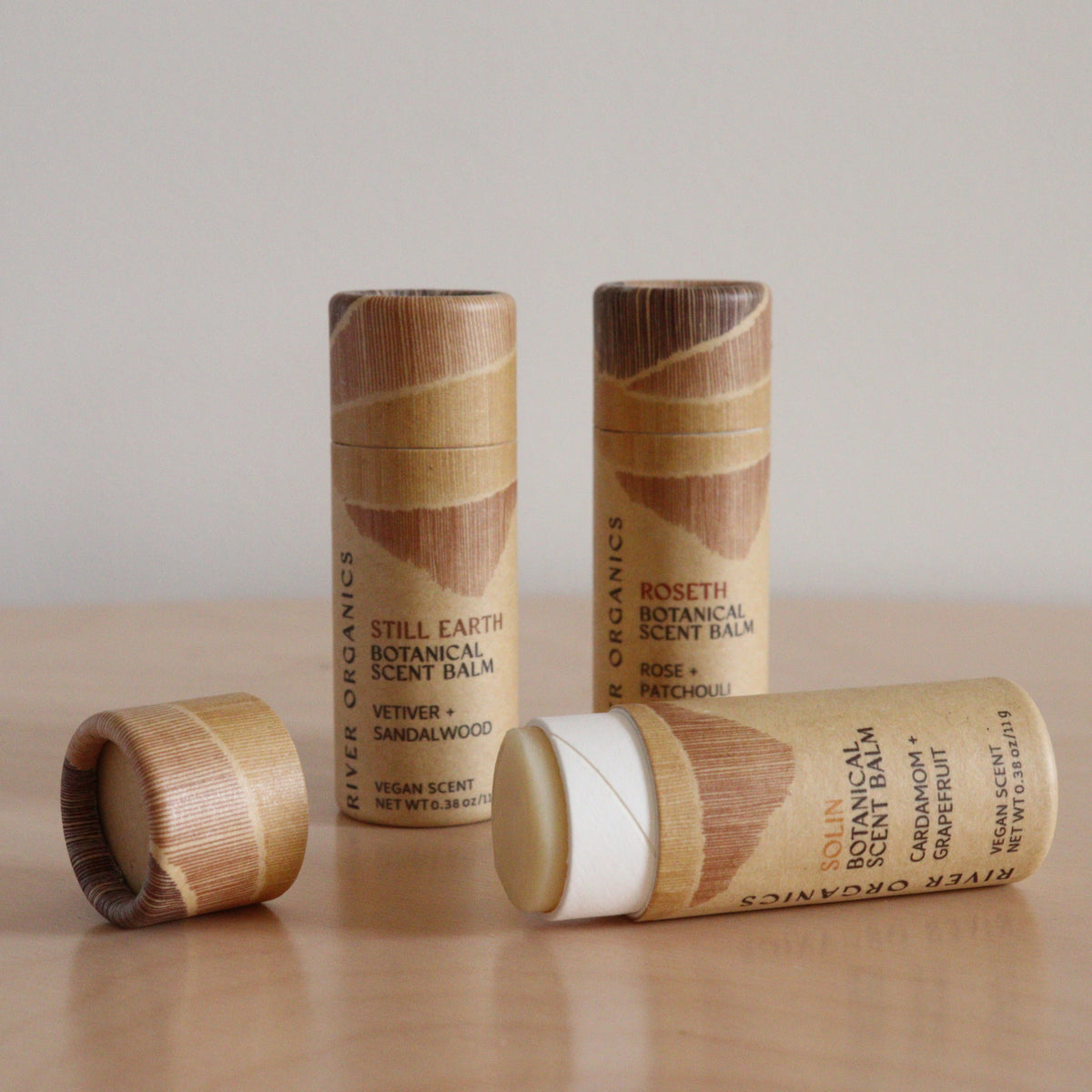 Three River Organics Botanical Scent Balms with essential oils are displayed on wood; one is open, showing the white balm inside, while two remain closed. The eco-friendly packaging highlights plant-based skincare and natural scents.