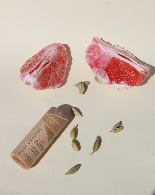 Two grapefruit wedges, several green cardamom pods, and Solin: Cardamom + Grapefruit Botanical Scent Balm by River Organics are arranged on a light cream background.