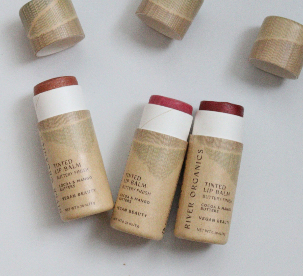 Three tubes of River Organics Tinted Vegan Lip Balm are displayed with their brown paper packaging and lids removed, revealing the lip balms in assorted shades. The labels emphasize cocoa butter and mango butter ingredients, as well as their vegan beauty credentials, making these organic, plastic-free lip balms an ideal choice for eco-conscious beauty enthusiasts.
