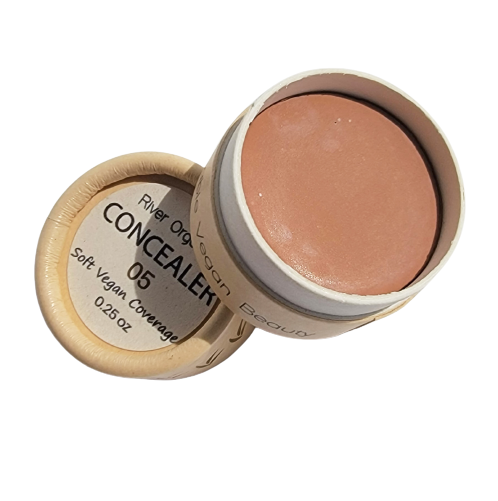 An open container of River Organics Imperfect Zero-Waste Vegan Concealer (Titanium dioxide-free) reveals the light brown cream inside. The lid label reads "Soft Vegan Coverage 0.26 oz." This gentle concealer offers an eco-friendly, plant-based solution.