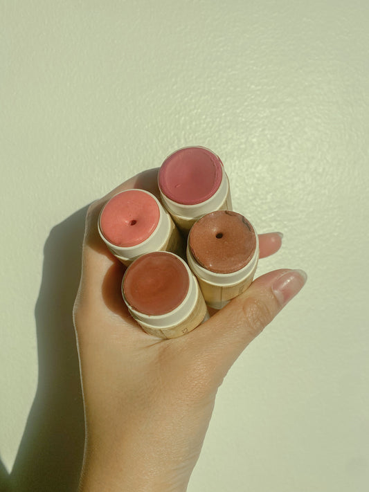 A hand holds four River Organics Natural Cream Blush Sticks—including a zero-waste option—in light pink, rosy pink, peach, and warm brown shades against a plain, light background.
