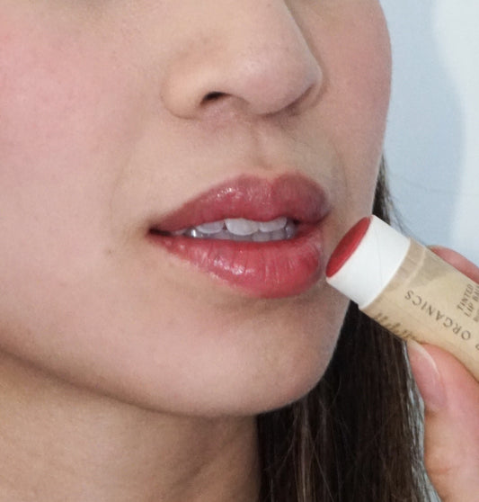 A person applies River Organics Tinted Vegan Lip Balm to their lips, holding the balm close to their mouth. The focus is on the lower half of the face.