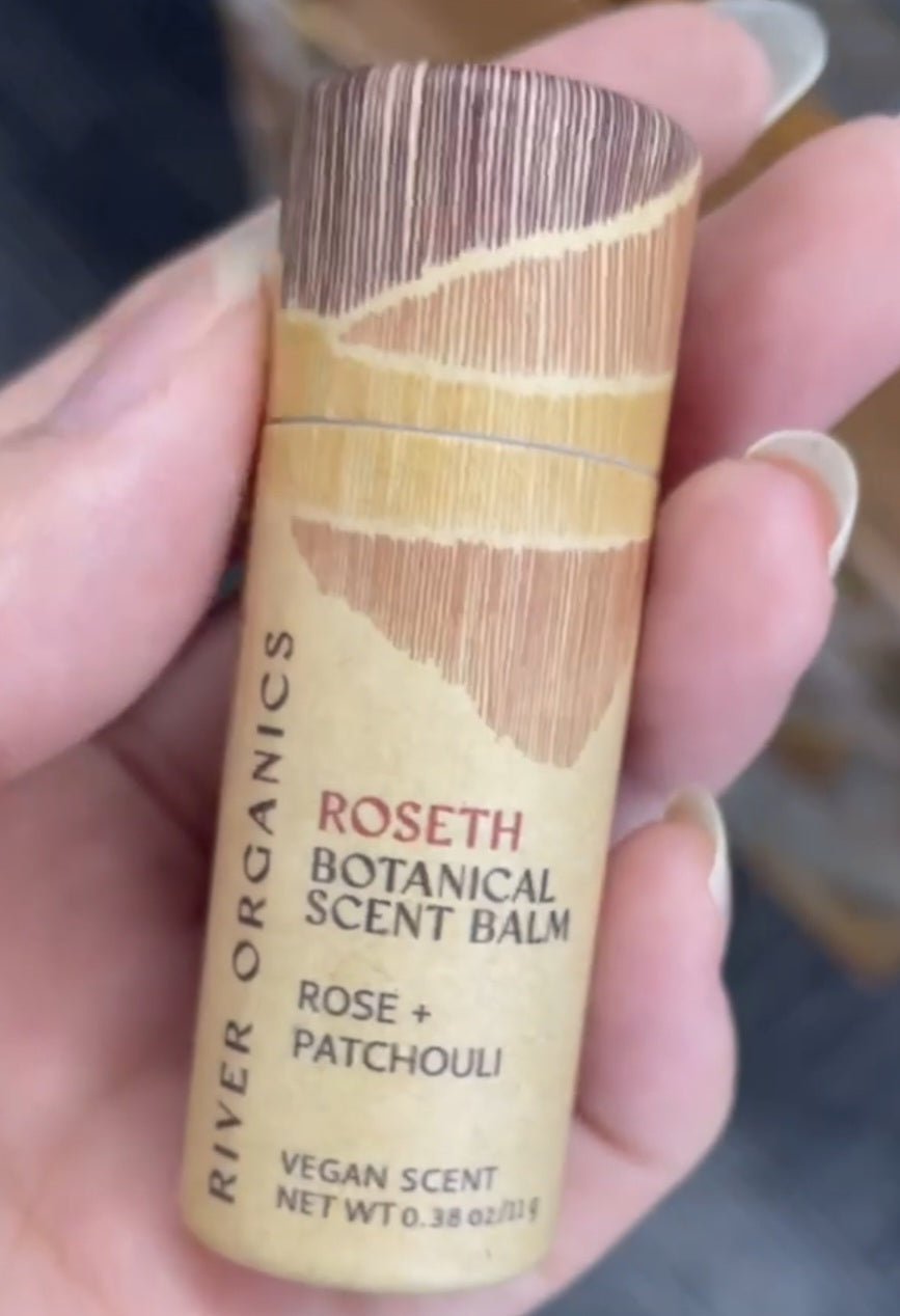 A hand holds River Organics' Roseth: Rose + Patchouli Botanical Scent Balm, a 0.38 oz vegan solid perfume with earthy-toned, abstract packaging and notes of rose and patchouli.