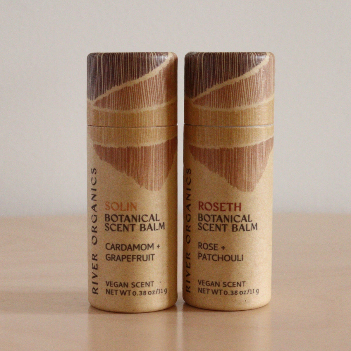 Two River Organics Botanical Scent Balms with brown, wood-like lids stand side by side: “Solin” (cardamom + grapefruit) and “Roseth” (rose + patchouli); both are solid perfumes made with essential oils.