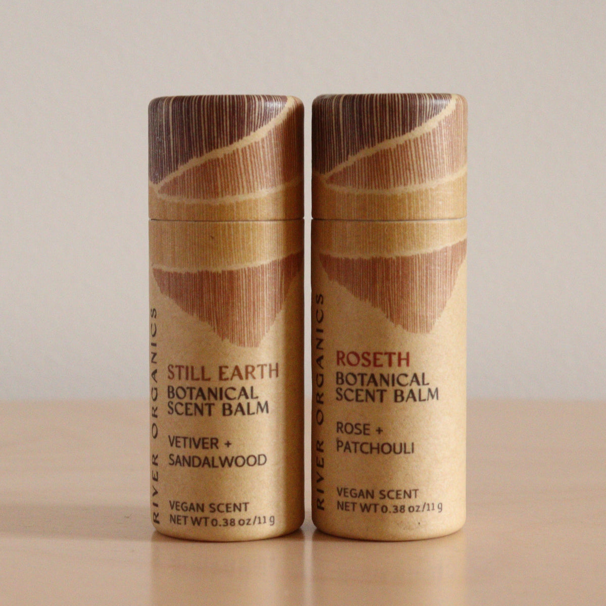 Two River Organics Botanical Scent Balms, made with essential oils, stand on a light wooden surface. The left tube says "Still Earth," the right says "Roseth." Both come in simple brown cylindrical packaging. Set of 2, 3 or 4 available.