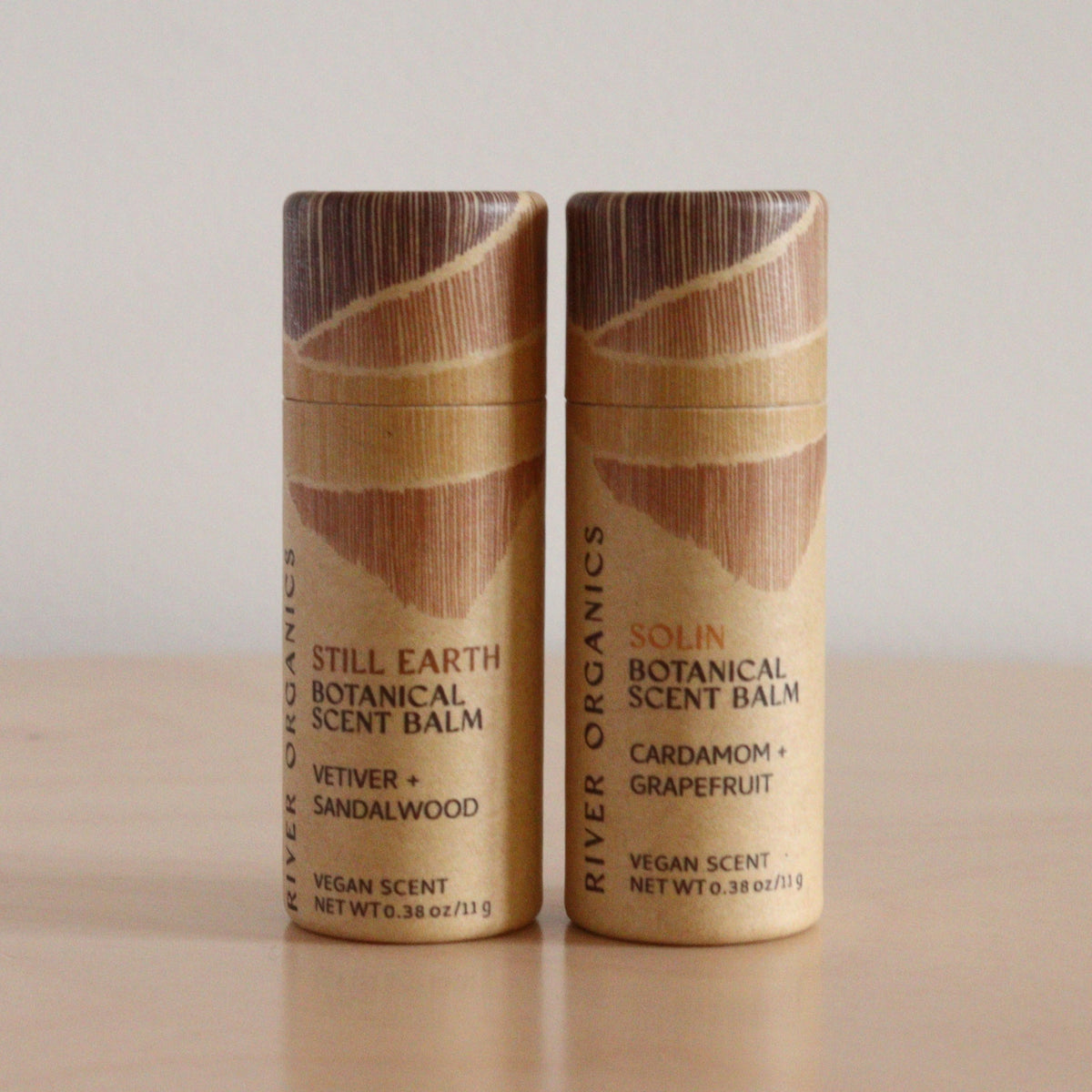 Two River Organics Botanical Scent Balm tubes (Set of 2) made with essential oils, in "Still Earth" (Vetiver + Sandalwood) and "Solin" (Cardamom + Grapefruit), stand on a light wooden surface.