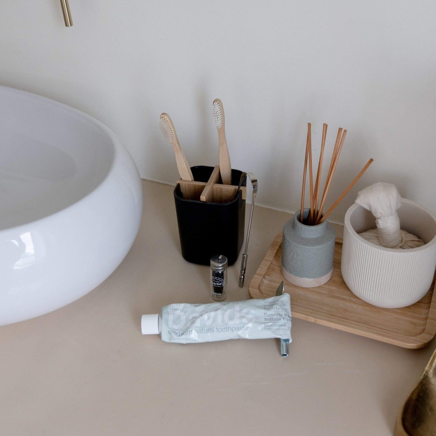 A bathroom countertop with a white sink, a black cup holding two bamboo toothbrushes and a razor, toothpaste, Bamboo Charcoal Floss by ZeroWasteStore.com, a small container, and a tray with a diffuser and rolled towel.