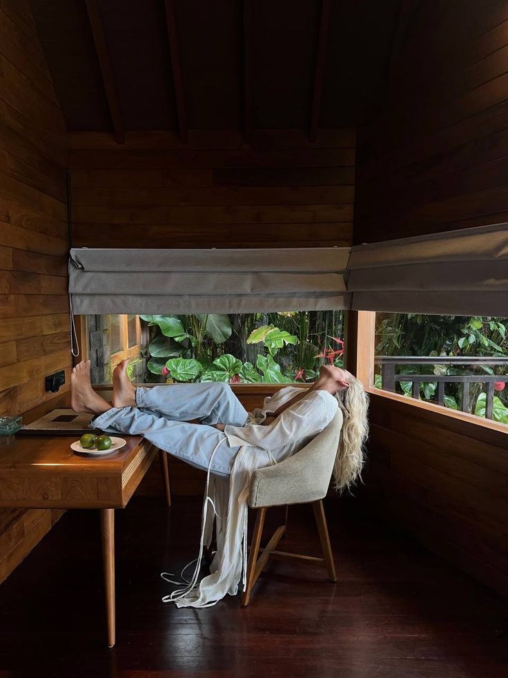 A person with long blond hair relaxes in a chair, feet up on a wooden desk in a cozy room filled with the calming aroma of River Organics’ Solin: Cardamom + Grapefruit Botanical Scent Balm, as large windows reveal lush greenery outside.