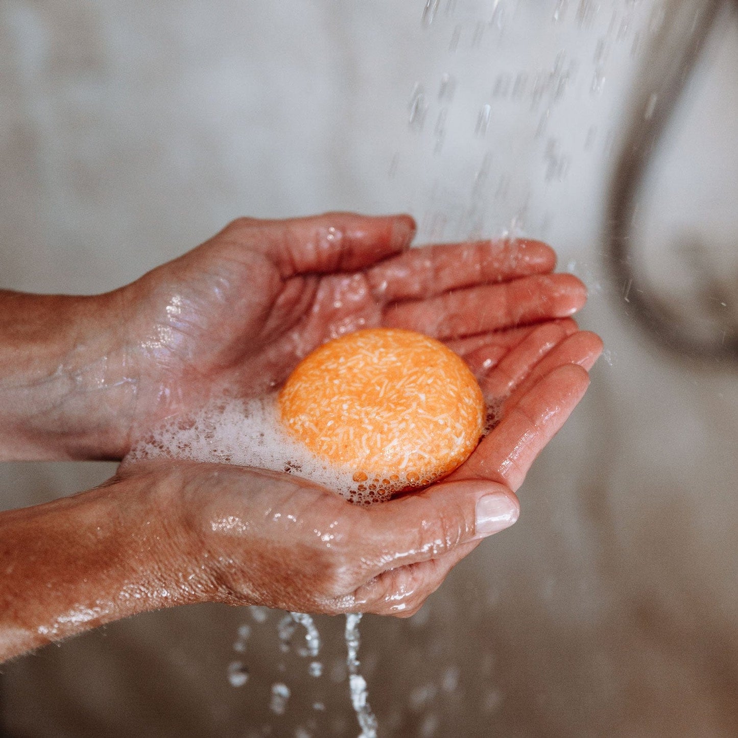 A person holds an orange Shampoo Bar from ZeroWasteStore.com, lathered with suds, under running water with both hands cupped together.