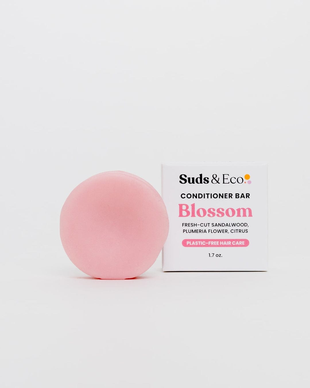 A round pink vegan Conditioner Bar by ZeroWasteStore.com sits next to its white box labeled "Conditioner Bar Blossom," showcasing its floral scent and zero waste, plastic-free hair care benefits, 1.7 oz., on a plain white background.