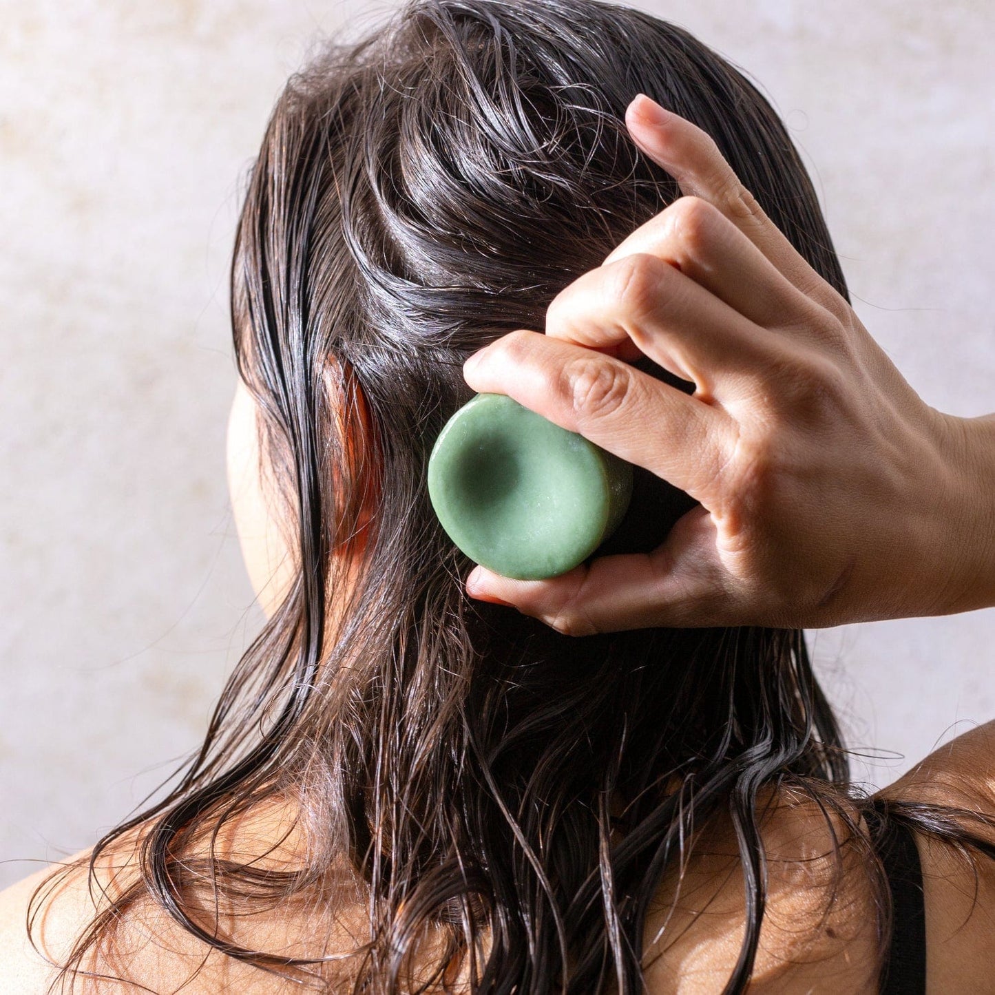 A person with wet hair is seen from behind, rubbing the green ZeroWasteStore.com Conditioner Bar onto the back of their head.