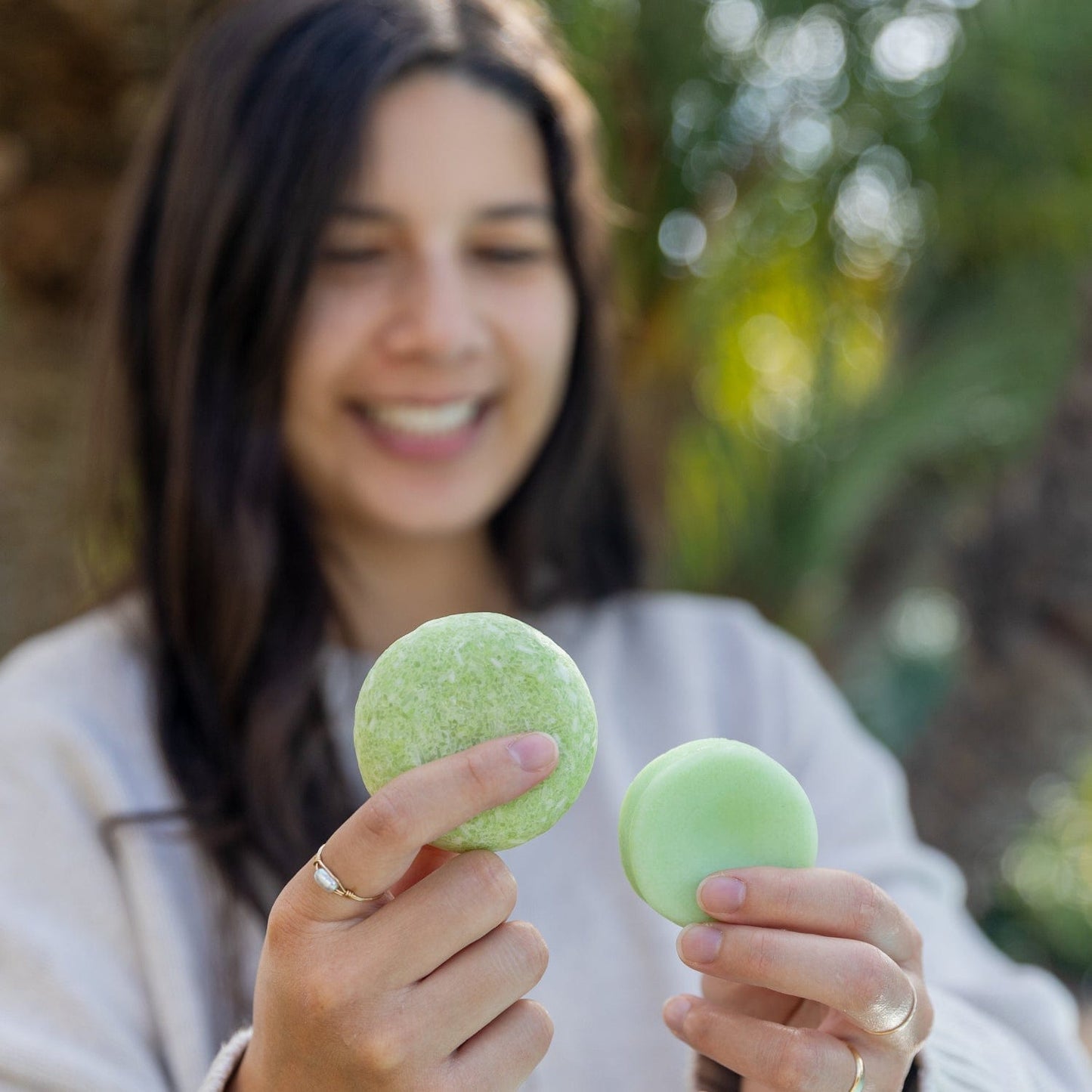A woman with long dark hair smiles outdoors amid lush greenery, holding a green vegan shampoo bar in one hand and a ZeroWasteStore.com Conditioner Bar container in the other.