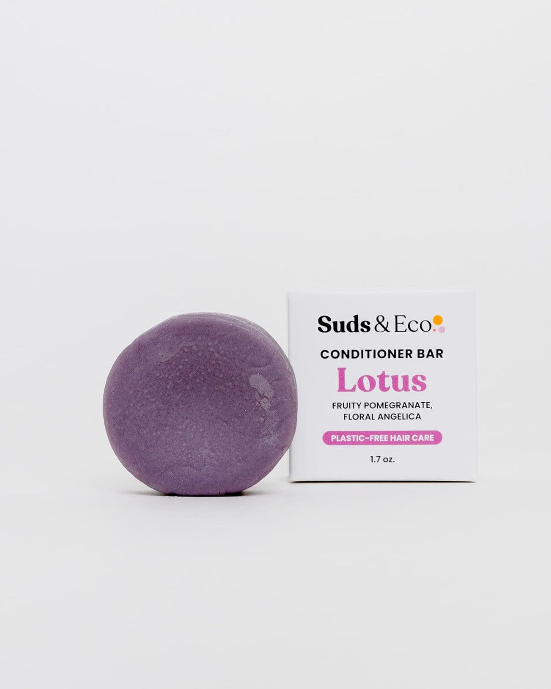 A round, purple vegan Conditioner Bar from ZeroWasteStore.com sits beside a white box labeled “Conditioner Bar—Lotus, Fruity Pomegranate, Floral Angelica, Plastic-Free Hair Care, 1.7 oz.” The plain white backdrop highlights this zero waste hair care choice.