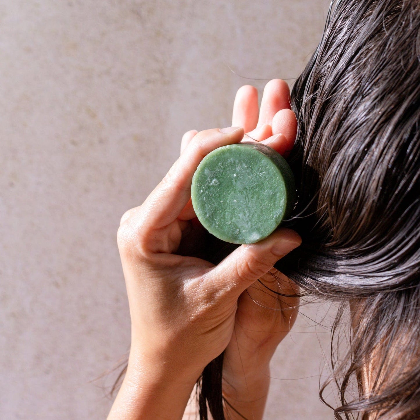 A person uses both hands to apply a green Conditioner Bar from ZeroWasteStore.com to their wet, dark hair, with a neutral background highlighting this eco-friendly, zero waste conditioning experience.