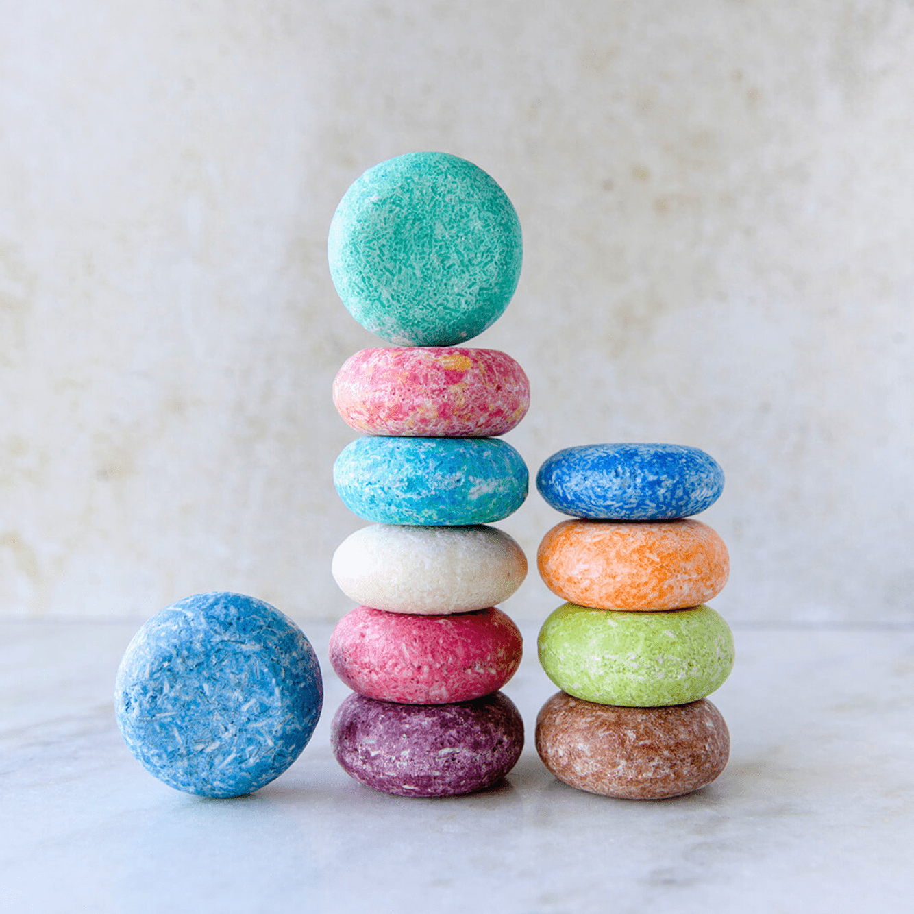 A stack of colorful, round Shampoo Bars from ZeroWasteStore.com sits on a light surface, featuring blue, pink, white, purple, brown, orange, and green bars for a vibrant zero waste haircare solution.