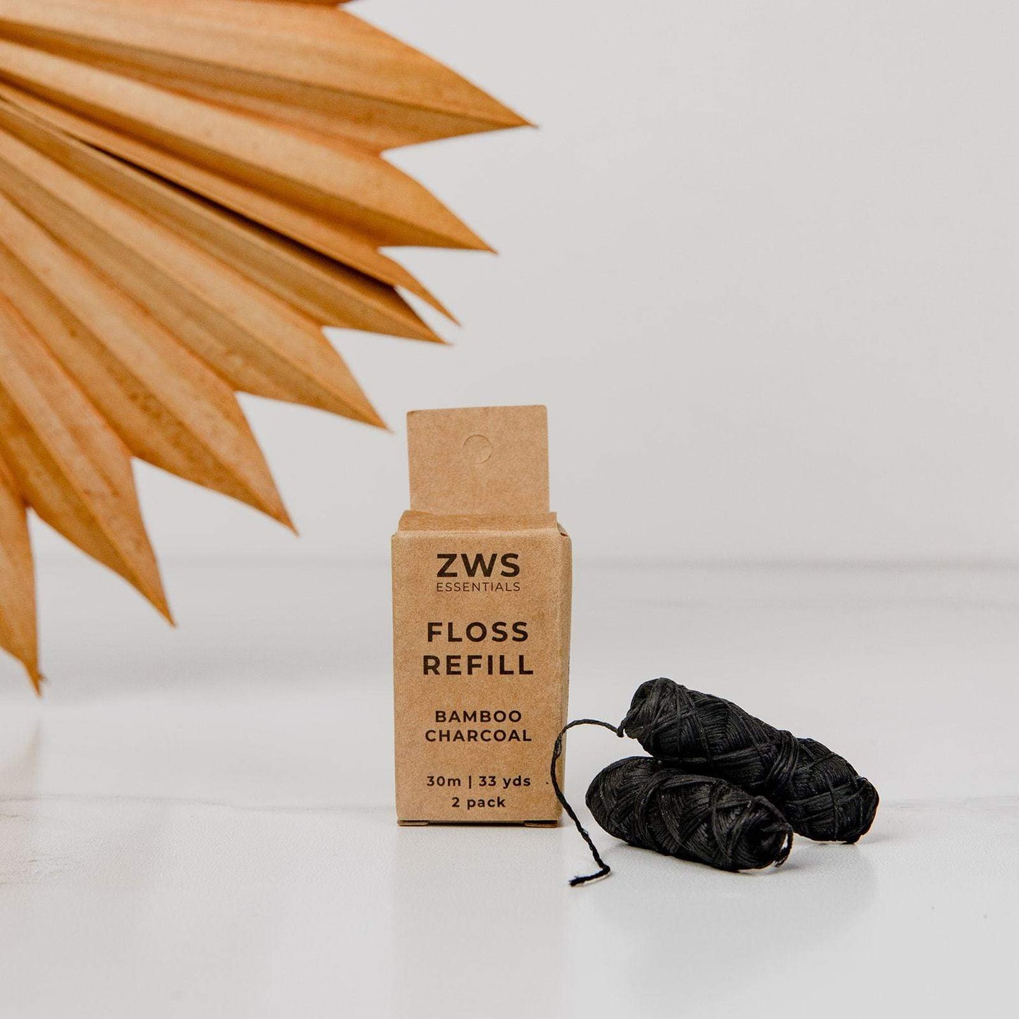 A ZeroWasteStore.com Bamboo Charcoal Floss box sits on a white surface beside two black biodegradable floss rolls, with a dried fan-shaped leaf in the background against a white wall.