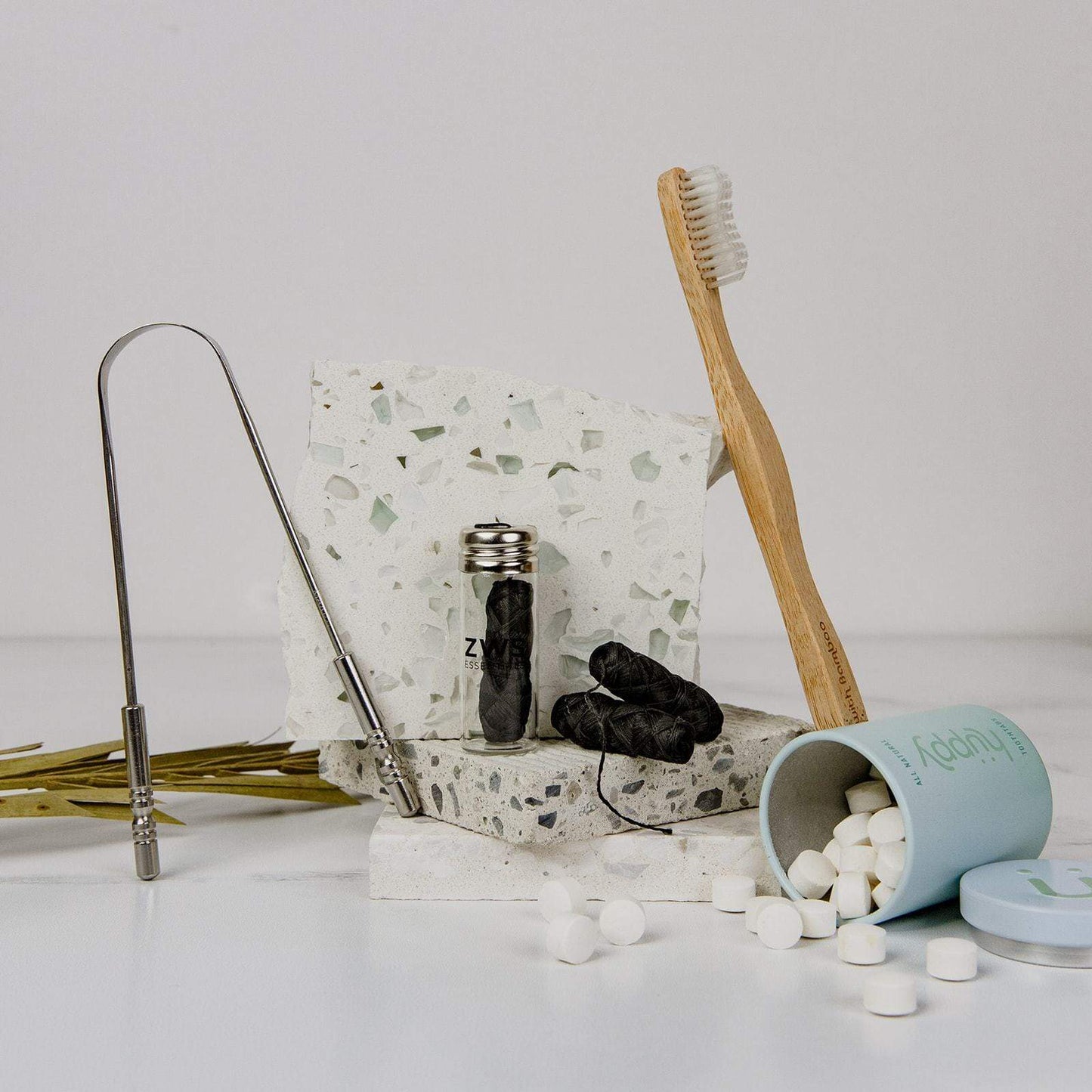 A set of eco-friendly dental care essentials from ZeroWasteStore.com, featuring a bamboo toothbrush, Bamboo Charcoal Floss in a glass jar, toothpaste tablets, and a metal tongue scraper displayed on terrazzo and white surfaces.
