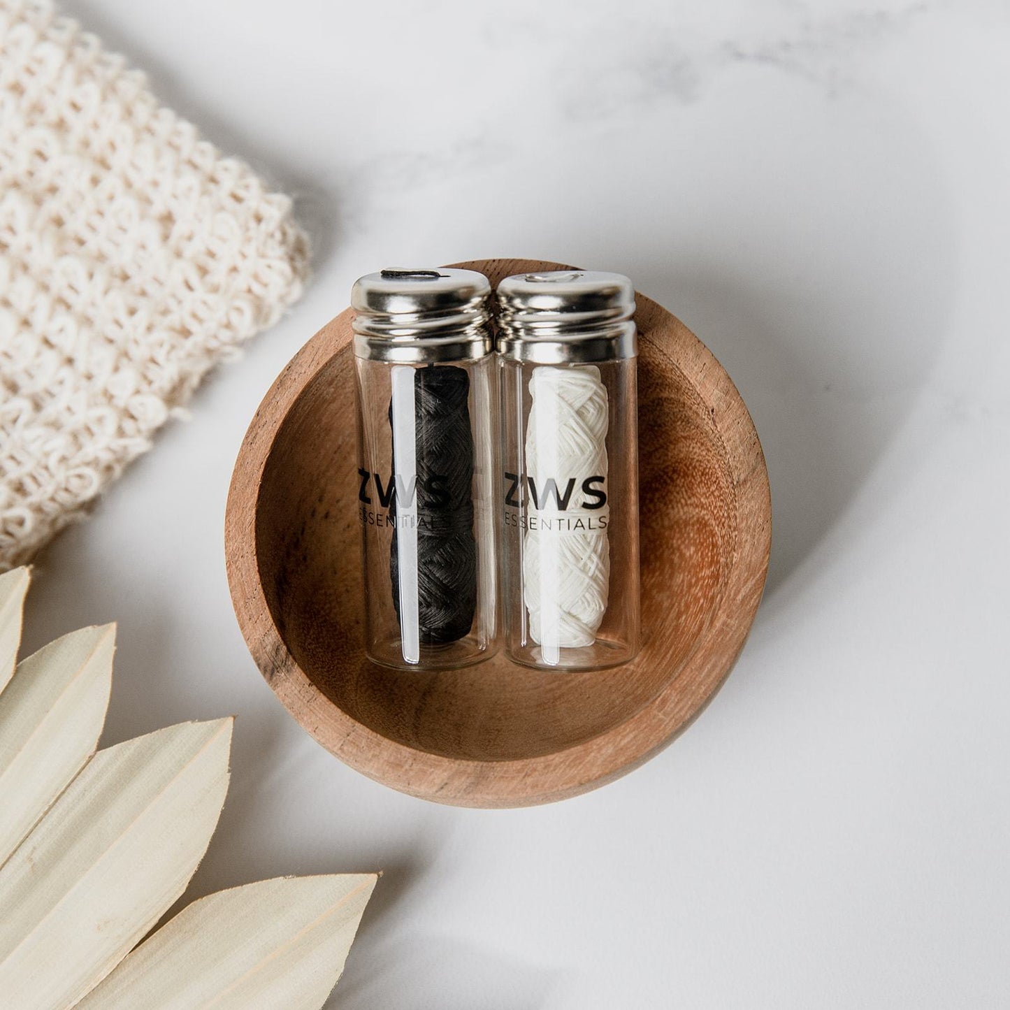 Two glass jars of ZeroWasteStore.com Bamboo Charcoal Floss—one black, one white—sit side by side with metal lids in a small wooden bowl on a light surface, next to knitted fabric and beige leaves.