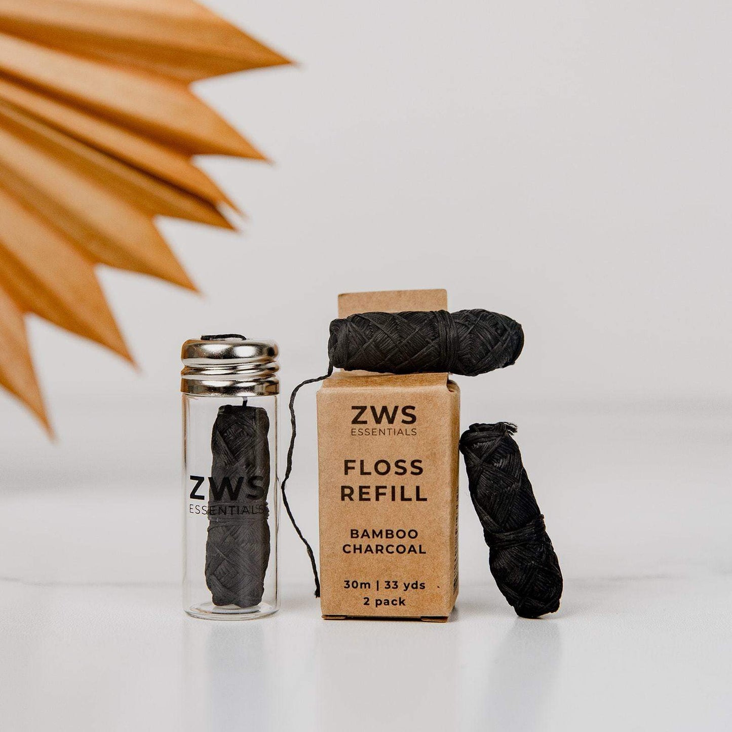 ZeroWasteStore.com’s Bamboo Charcoal Floss is shown in a glass jar, next to a "ZWS Essentials Floss Refill" box and two biodegradable floss spools, all arranged on a white surface with a brown leaf in the background.