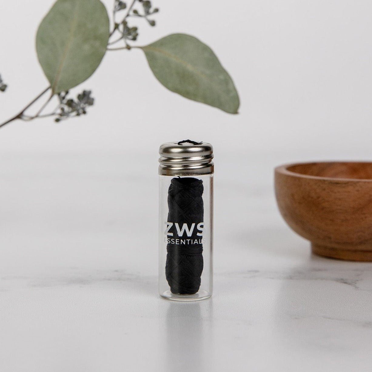 A small glass jar with a metal lid containing Bamboo Charcoal Floss from ZeroWasteStore.com rests on a white surface, with a wooden bowl and eucalyptus leaves in the background.