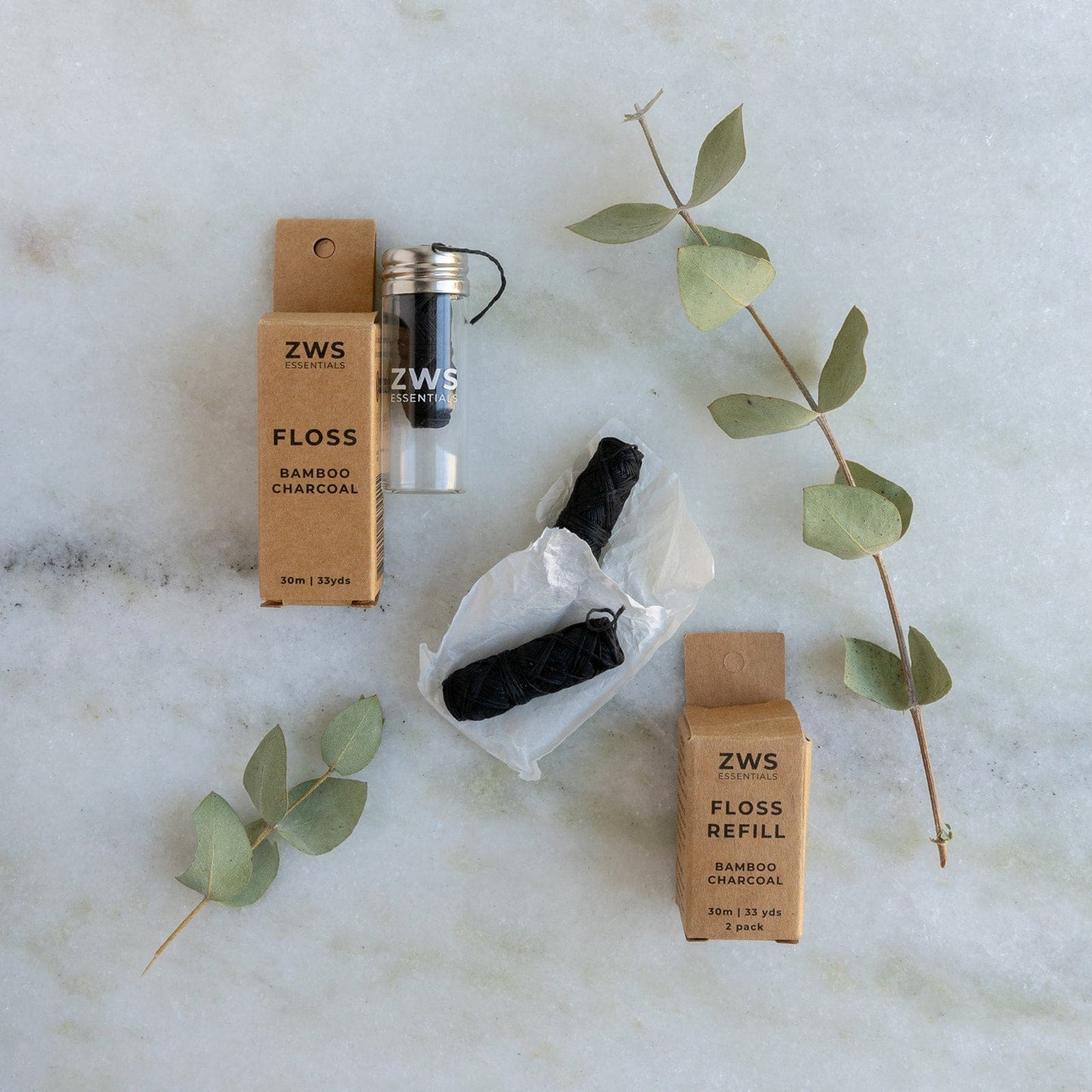 ZeroWasteStore.com's Bamboo Charcoal Floss, with a glass container and two biodegradable refills in zero-waste cardboard packaging, is displayed on light marble with eucalyptus sprigs nearby.