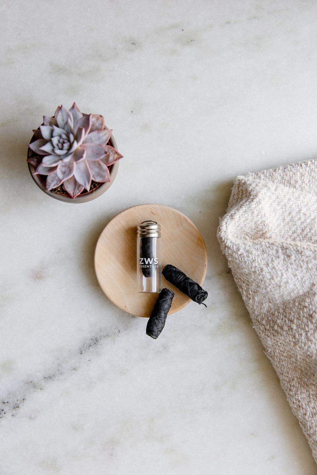 A small succulent in a pot sits on a marble surface beside a wooden dish with a glass container labeled “ZW,” two black items, Bamboo Charcoal Floss by ZeroWasteStore.com, and a beige textured towel.