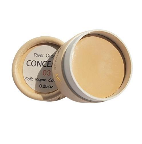 A small, round container of River Organics Zero-Waste Vegan Concealer with the lid off shows a creamy beige product. The label reads "Zero-Waste Vegan Concealer 0.25 oz.