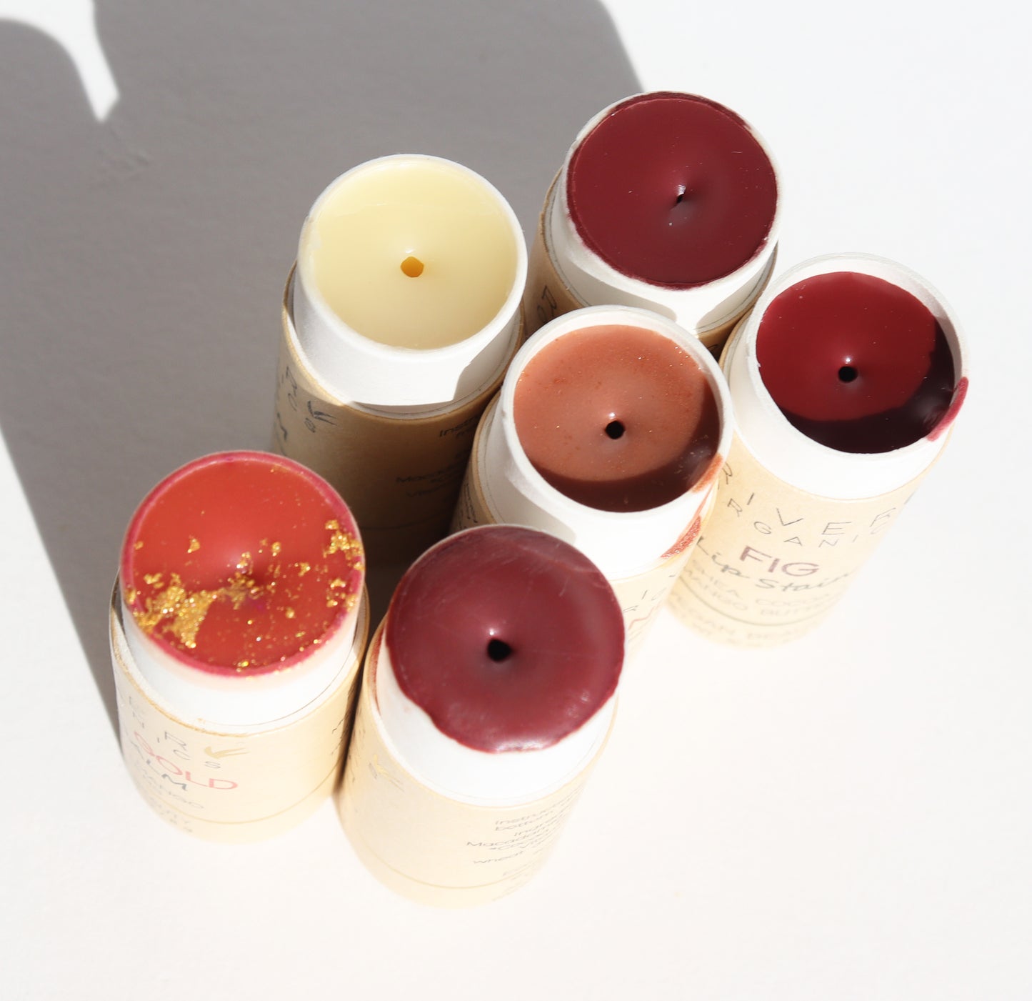 Six tubes of River Organics Imperfect Lip Balms + Lip Stains sit on a white surface, showcasing shades of red, pink, and beige. Some feature visible gold glitter. The partially opened tubes reveal the balms inside while they cast shadows under a strong light source.