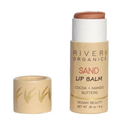 A tube of River Organics Tinted Vegan Lip Balm with the cap off reveals the balm. The beige and brown packaging, with a leaf design, reads "Cocoa + Mango Butters" and "Vegan Beauty," highlighting deep moisture from plant-based ingredients.