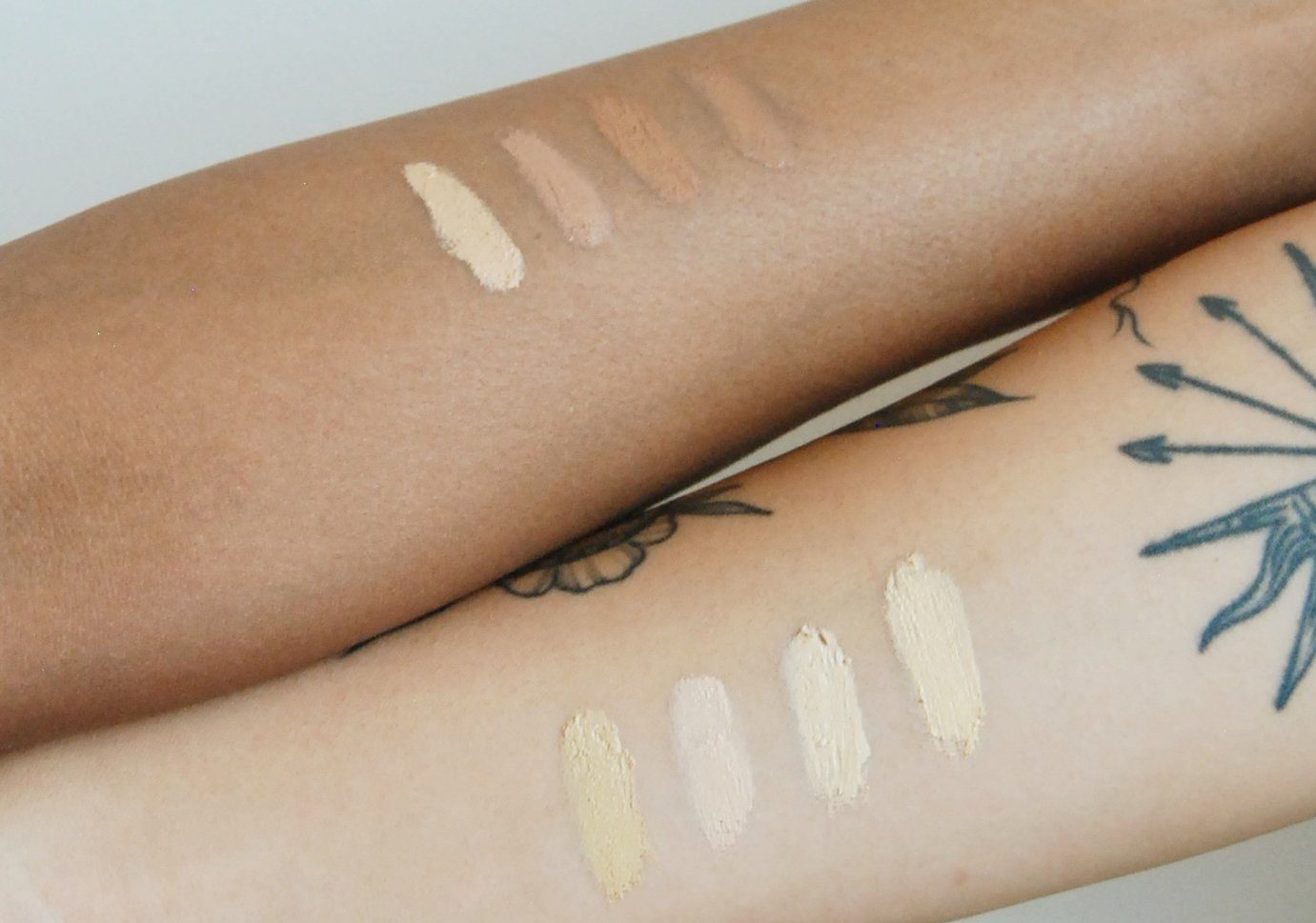 Two forearms—one lighter with tattoos, one darker—show swatches of River Organics Zero-Waste Vegan Concealer in various beige and brown tones; the lighter arm has four swatches, the darker has five.