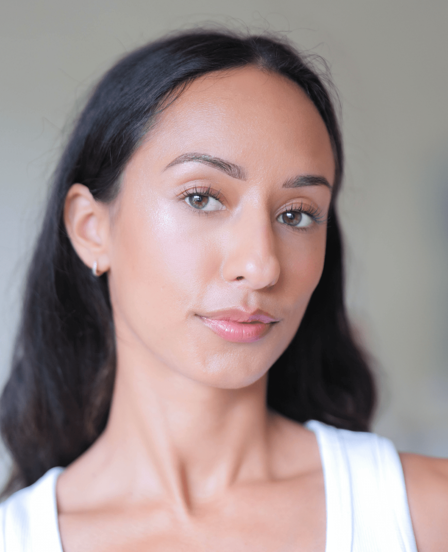 A woman with long dark hair and glowing skin looks at the camera, wearing a white top and small hoop earrings, against a blurred neutral background.