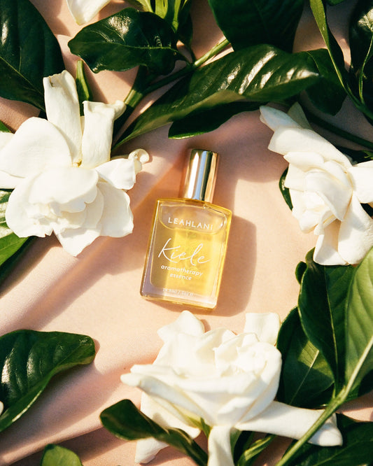 LEAHLANI Kiele Essence of Gardenia, adorned with a gold cap, is set among white florals and glossy green leaves on a soft pink surface, capturing the delicate beauty of gardenia perfume.