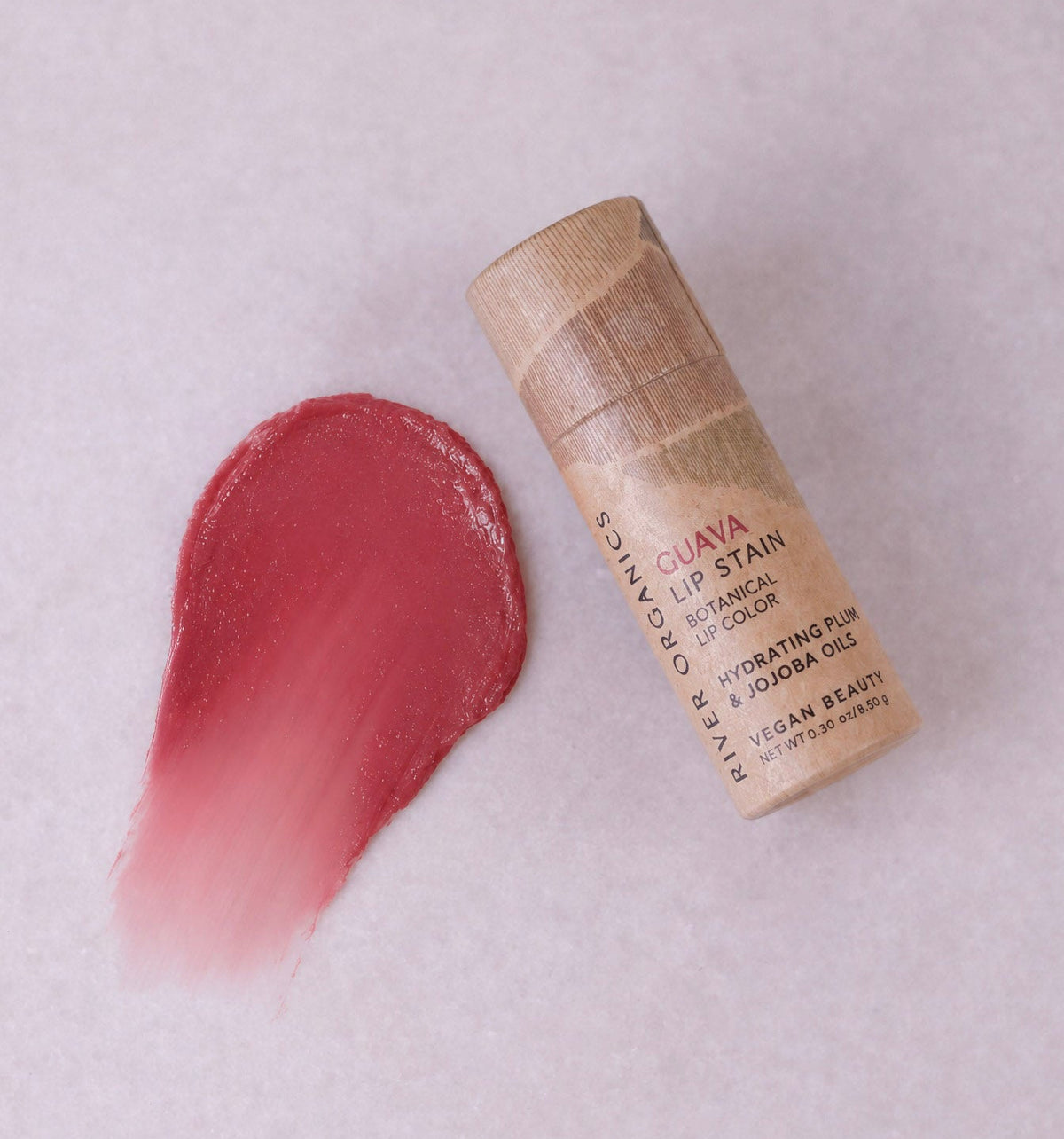 A tube of River Organics Lip Stain: Vegan, Plastic-Free, & Organic with a wooden cap lies next to a swatch of the pink shade on a light surface, highlighting this eco-friendly lip stain’s sustainable design and vibrant color.