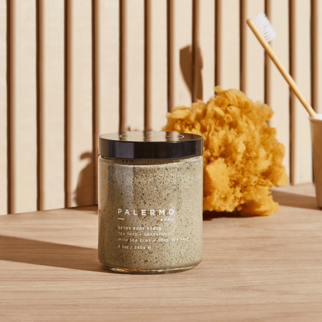 A jar of Palermo Body Detox Body Scrub with a black lid sits on a beige surface. Behind it, a yellow natural sponge and a toothbrush in a cup are set against a wooden panel wall. The scrub contains mineral-rich Dead Sea salt.