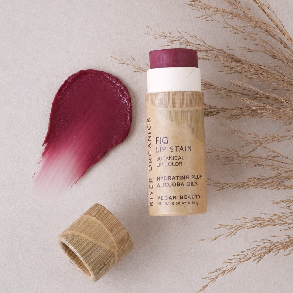 A tube of River Organics Lip Stain: Vegan, Plastic-Free, & Organic | All Colors, with its cap off, rests next to a swatch of the lip color on a beige surface, accented by dried grass as decoration.