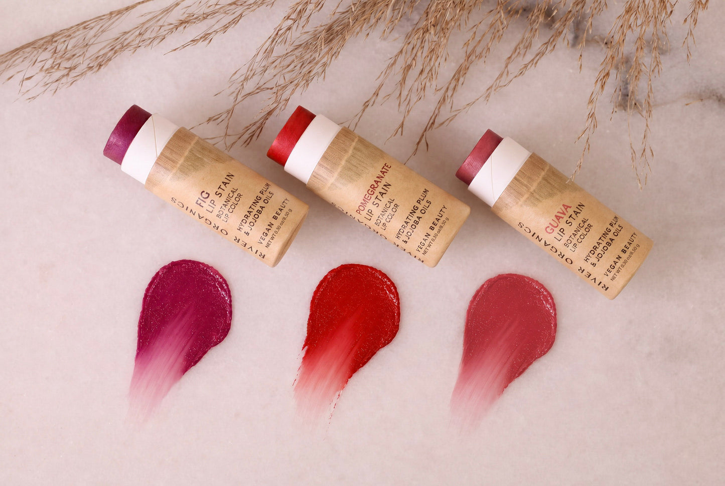 Three tubes of River Organics Lip Stain: Vegan, Plastic-Free, & Organic | All Colors are arranged on a white surface, each accompanied by a swipe of deep pink, red, or rose beneath them. Sprigs of dried grass accent the top of the scene, underscoring the natural elegance of this eco-friendly lip stain collection.