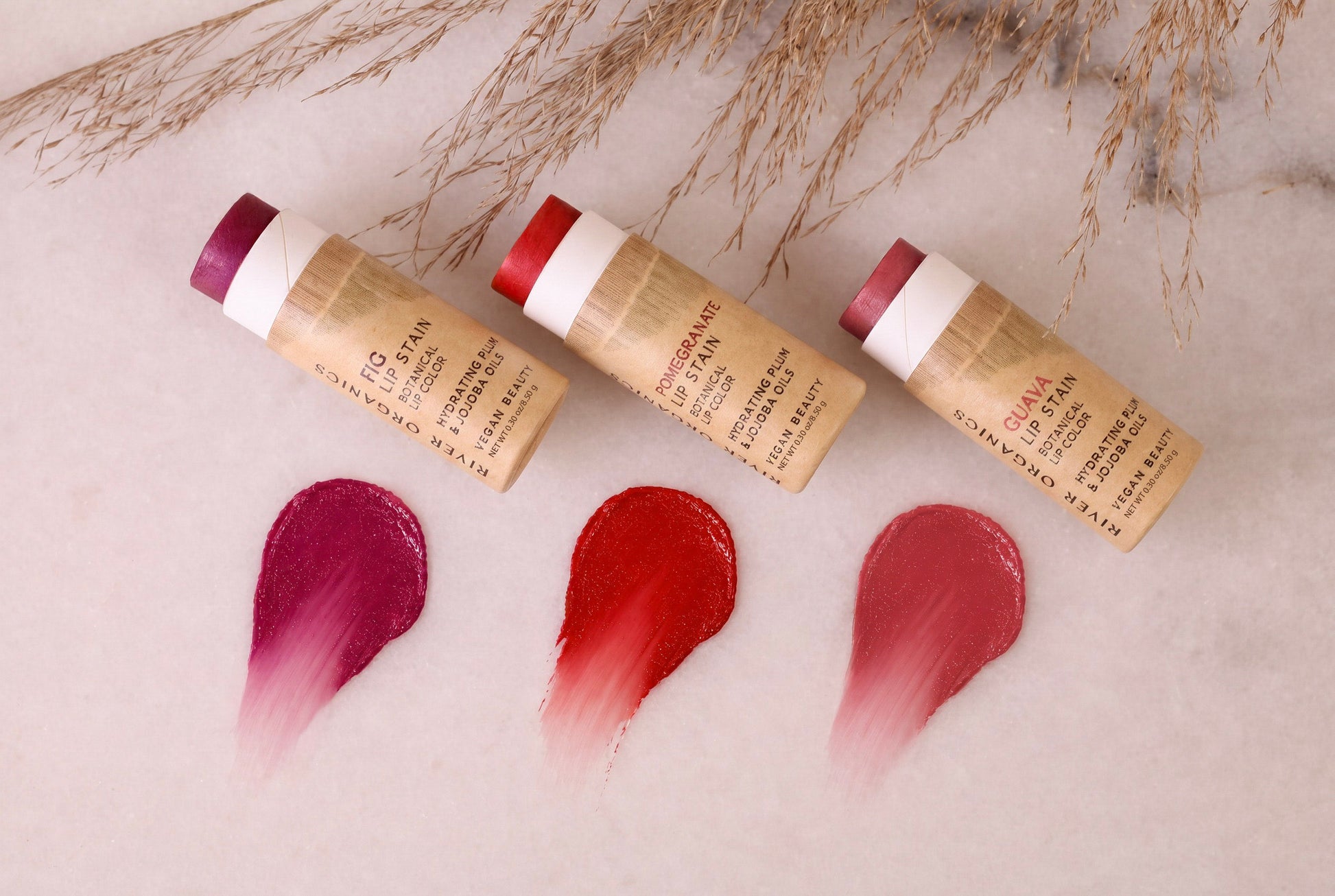 Three tubes of River Organics Lip Stain: Vegan, Plastic-Free, & Organic | All Colors are arranged on a white surface, each accompanied by a swipe of deep pink, red, or rose beneath them. Sprigs of dried grass accent the top of the scene, underscoring the natural elegance of this eco-friendly lip stain collection.