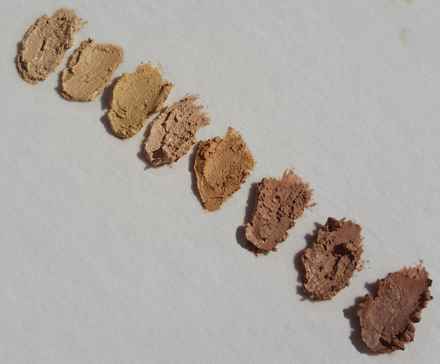 Eight swatches of River Organics' Imperfect Natural Hydrating Vegan Concealer, in shades from light beige to deep brown, are smeared diagonally on white, showcasing skin tones with organic plant oils and zero-waste formulas.