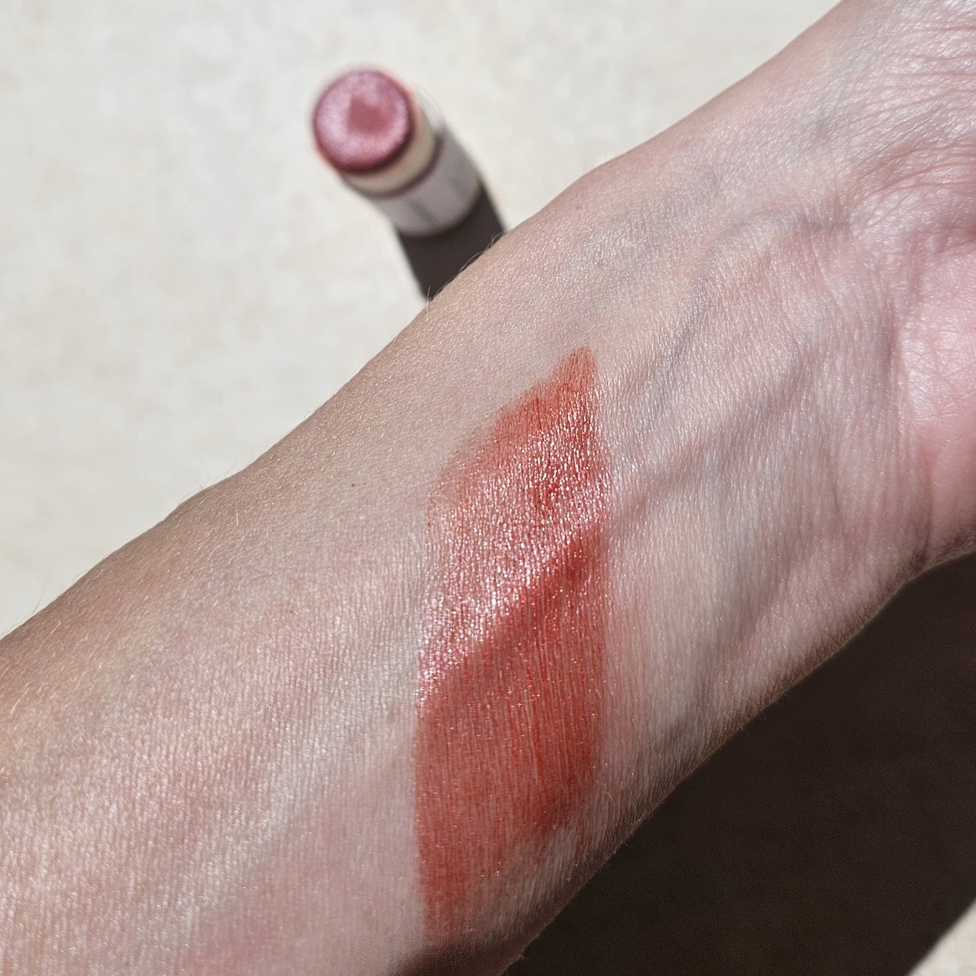 A swatch of shimmery, reddish-brown Pomegranate Vegan Lip Stain by River Organics is displayed on the inner side of a person's wrist. In the background, the lipstick tube with eco-friendly packaging is visible with its cap off. The image is taken under natural light on a light-colored surface.