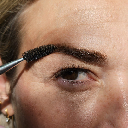An eyebrow spoolie brush applying dark tinted River Organics Brow Wax & Growth Serum - All Colors to an eyebrow.