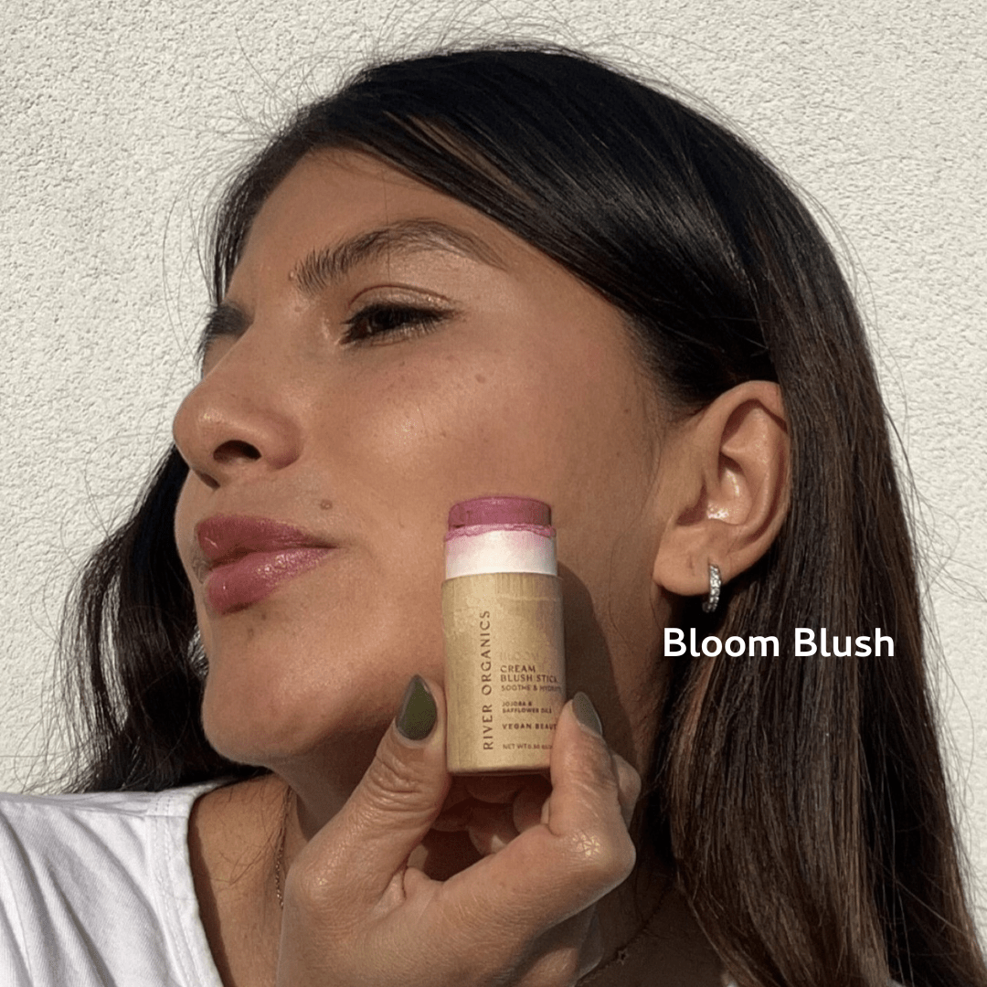 A woman applies River Organics Natural Cream Blush Stick to her cheek, highlighting a fresh makeup look. "Bloom Blush" appears by her face, emphasizing its plastic-free packaging against a light background.