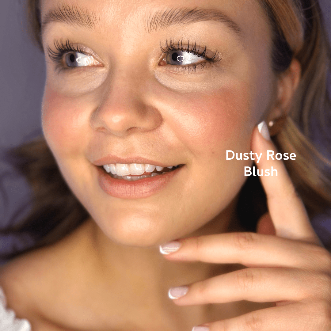 A woman with glowing skin softly smiles, showcasing River Organics Natural Cream Blush Stick in a rosy shade on her cheeks. "Natural Cream Blush Stick" appears near her cheek as she wears manicured nails and a white top against a neutral backdrop.