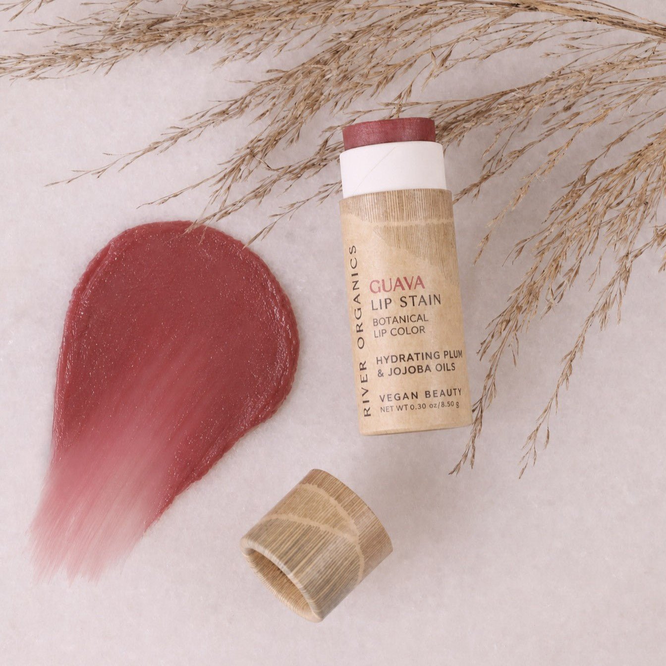 An open tube of River Organics Lip Stain: Vegan, Plastic-Free, & Organic | All Colors rests beside a pink swipe of lip color and dried grasses on a light backdrop. The label highlights nourishing plum and jojoba oils, presenting this long-lasting hydrating formula as an organic, plastic-free vegan lip stain—ideal for the conscious beauty enthusiast.