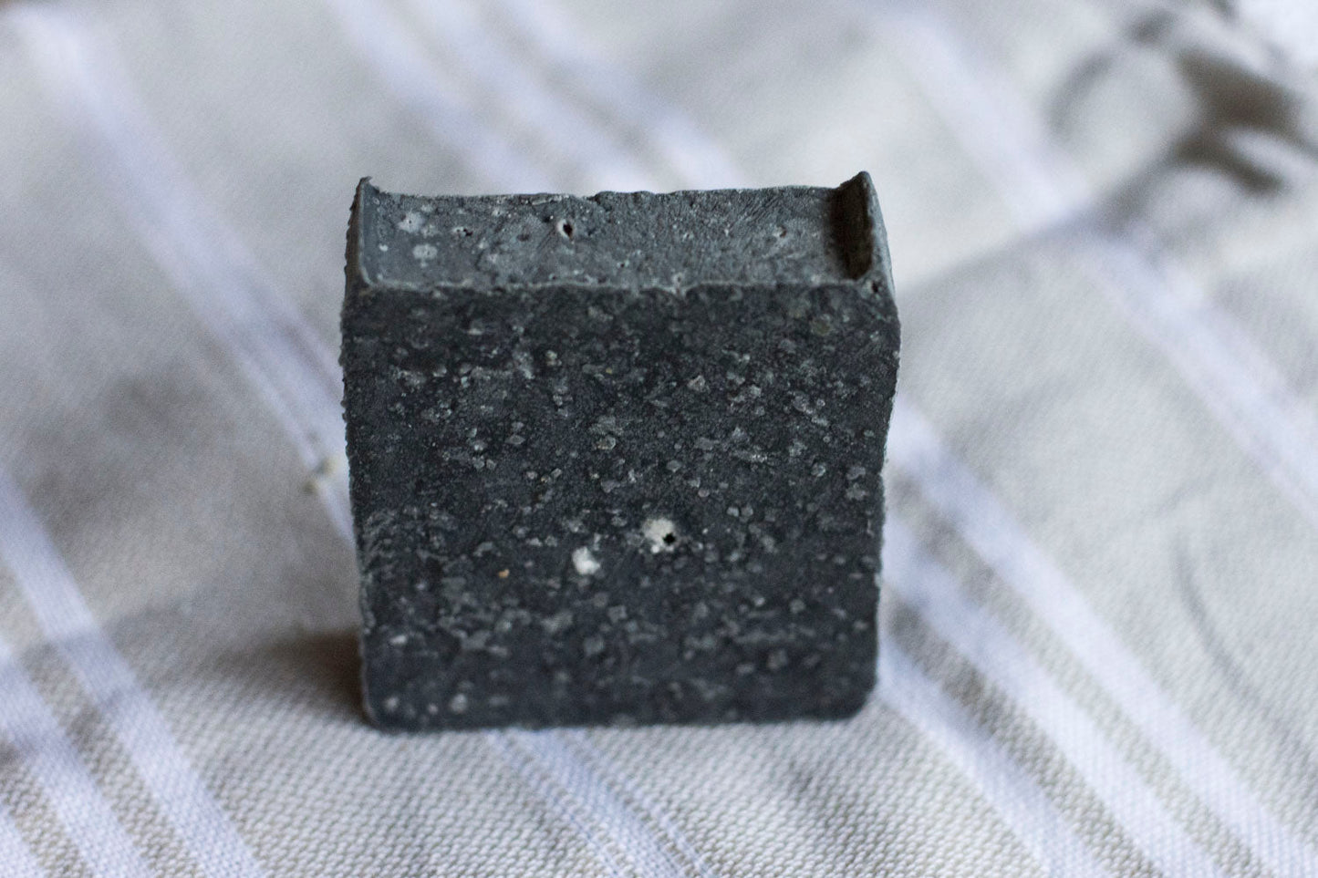 The SOLIDSILK® ONYX Facial Cleansing Bar by NO TOX LIFE is a small, black, square bar with a rough, speckled texture. Made with activated charcoal, it offers a deep clean and sits upright on a light-colored, striped fabric surface.